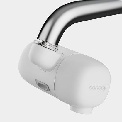The Canopy Faucet Filter, a sleek white device by Canopy, attaches to a curved metal faucet for water filtration. It features a modern design with a button or sensor on the front and is fulfilled by Canopy.
