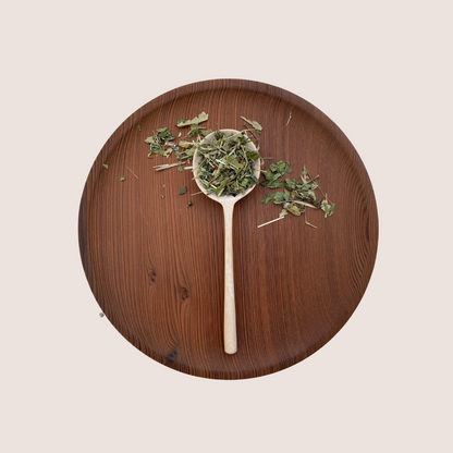 A wooden spoon filled with Moon Canyon Healing's Fill The Well Herbal Infusion rests on a wooden plate. The plate and spoon are both light brown, complementing the mixture of green and brown hues of the herbs that promise nourishment for your daily routine. The background is a plain, light beige.