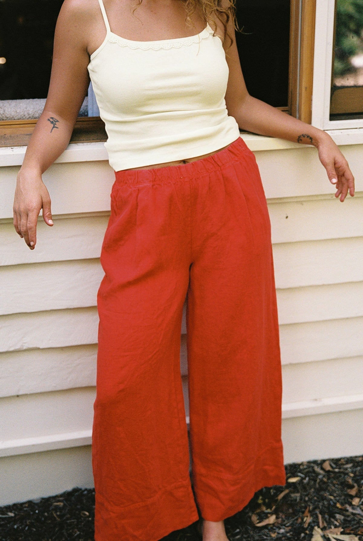 Cream - 
A person stands outside in front of a white wall wearing the LA RELAXED Frill Camisole in pale yellow and loose red pants. Their face is not visible, and one arm rests on a window frame.