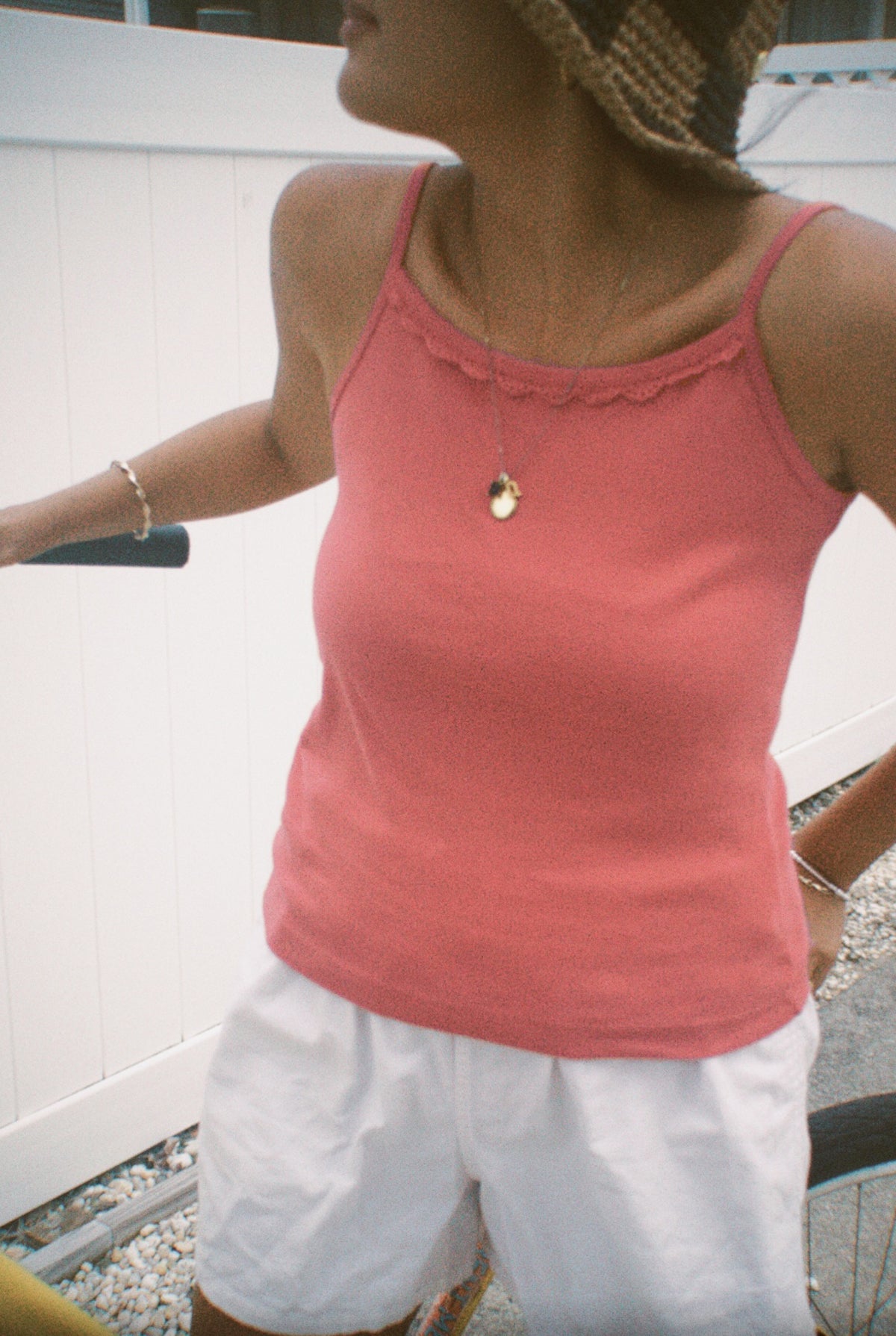 Rose - 
A woman wears the LA RELAXED Frill Camisole in pink with white shorts, a straw hat, and jewelry, standing by a bicycle near a white fence on gravel. Her face is partially out of frame.