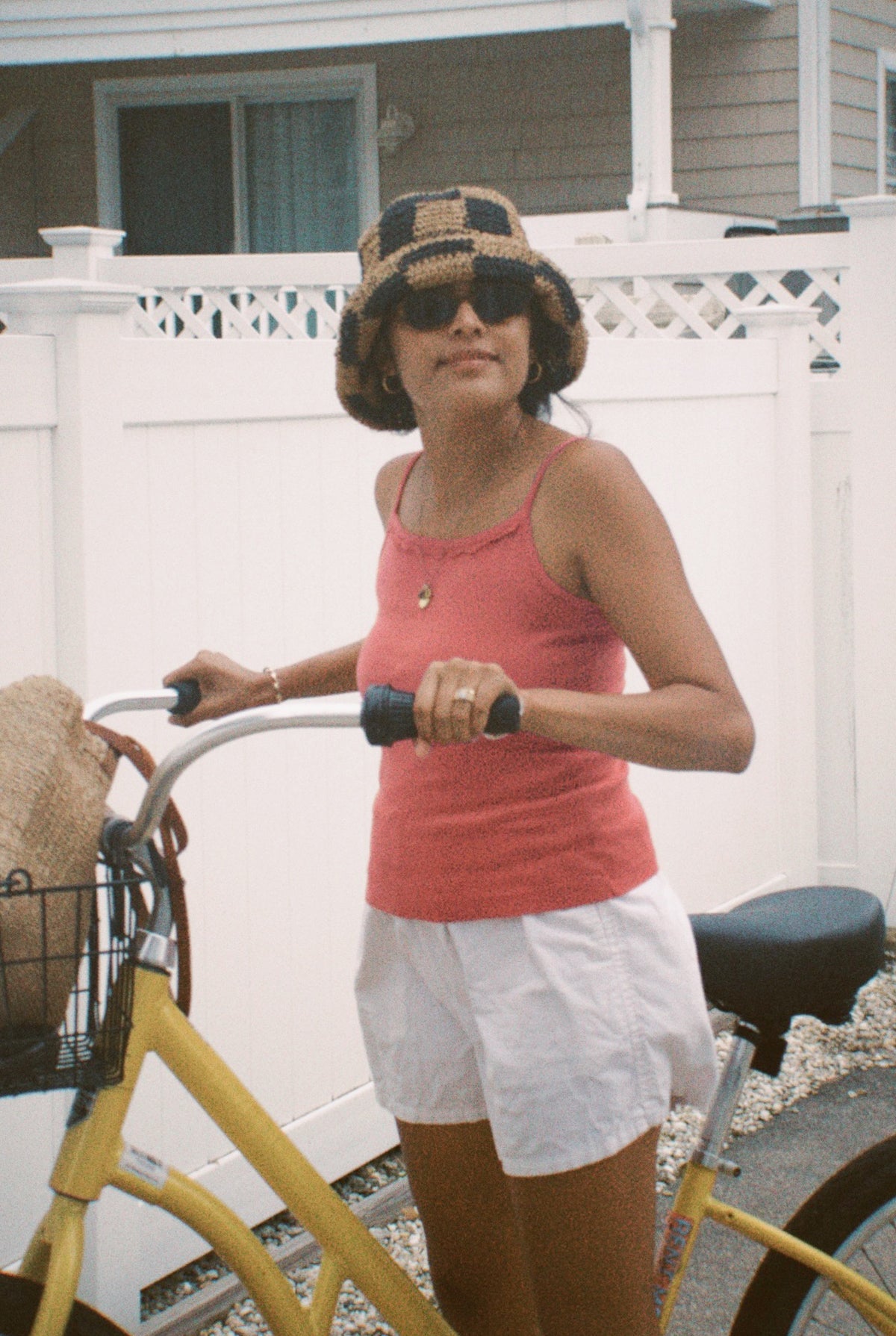 Rose - 
A woman wearing the LA RELAXED Frill Camisole, white shorts, sunglasses, and a checkered bucket hat holds a yellow bicycle with a woven bag in its basket, posing in front of a white fence and house.