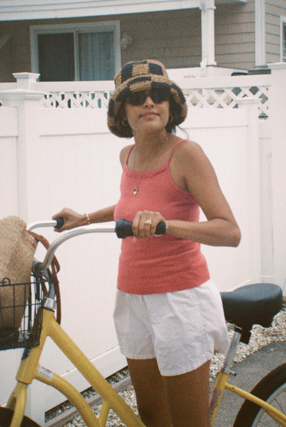 Rose - 
A woman wearing the LA RELAXED Frill Camisole, white shorts, sunglasses, and a checkered bucket hat holds a yellow bicycle with a woven bag in its basket, posing in front of a white fence and house.