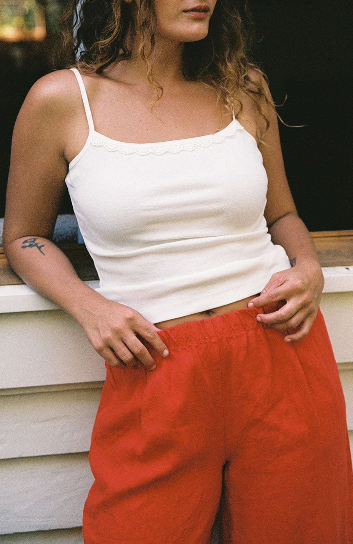 Cream - 
A woman with wavy hair wears the LA RELAXED Frill Camisole, a white lace-trim top, paired with loose red pants and stands in front of a white wall, her hands on her waistband. Only her upper body is visible.