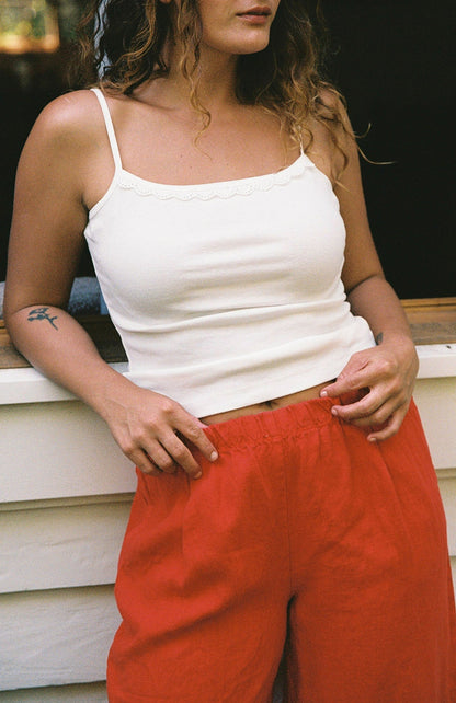 Cream - 
A woman with wavy hair wears the LA RELAXED Frill Camisole, a white lace-trim top, paired with loose red pants and stands in front of a white wall, her hands on her waistband. Only her upper body is visible.