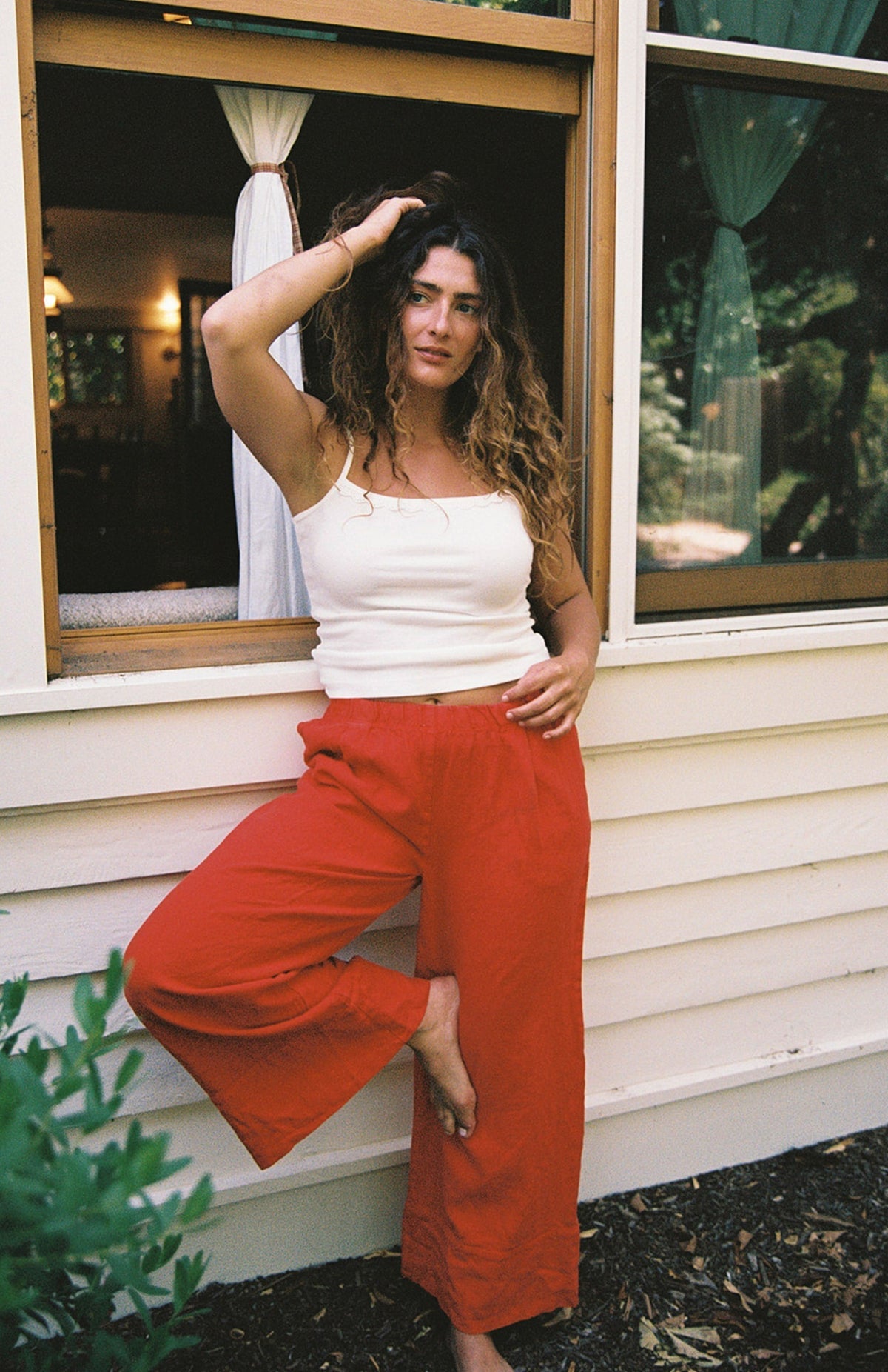 Cream - 
A woman with curly hair wears the LA RELAXED Frill Camisole and red pants, standing barefoot by an open window with one leg bent, her hand in her hair as she looks at the camera.