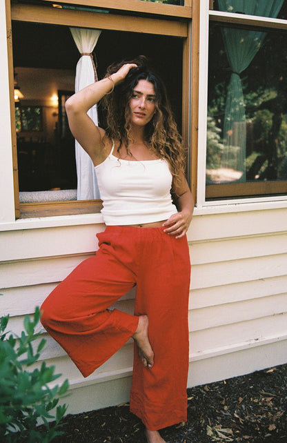 Cream - 
A woman with curly hair wears the LA RELAXED Frill Camisole and red pants, standing barefoot by an open window with one leg bent, her hand in her hair as she looks at the camera.