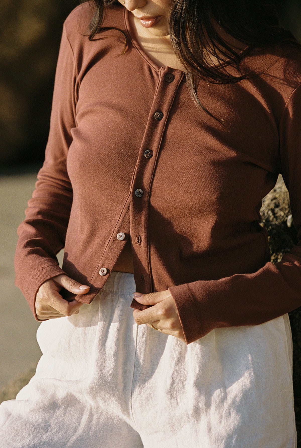 Mocha 
Someone is outdoors in natural sunlight, wearing an LA Relaxed Organic Rib Cardigan in rust over a white organic cotton top with white pants. They are adjusting the cardigan's buttons, creating a 90's closet vibe focused on the clothing.