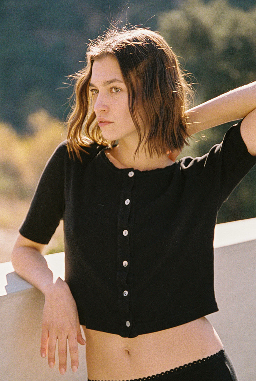 A person with shoulder-length hair stands outside, leaning on a ledge. They're wearing the Short Sleeve Organic Cardi by LA Relaxed, which features shell buttons. Their hand rests on their neck, and the blurred background reveals greenery and sunlight.