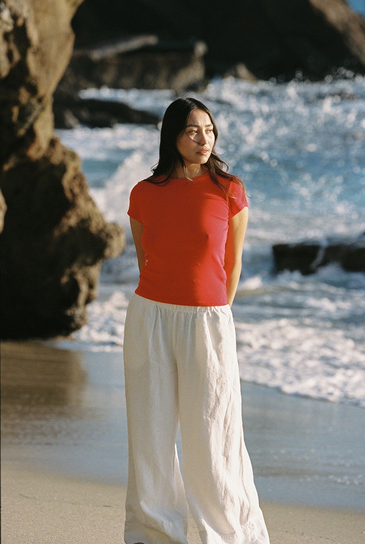 Cherry On a sandy beach near the ocean, someone wears an LA Relaxed Bebe Organic Tee and linen pants. Gentle waves crash, and rocky cliffs frame the scene as they enjoy the sunny day in a calm, relaxed manner.