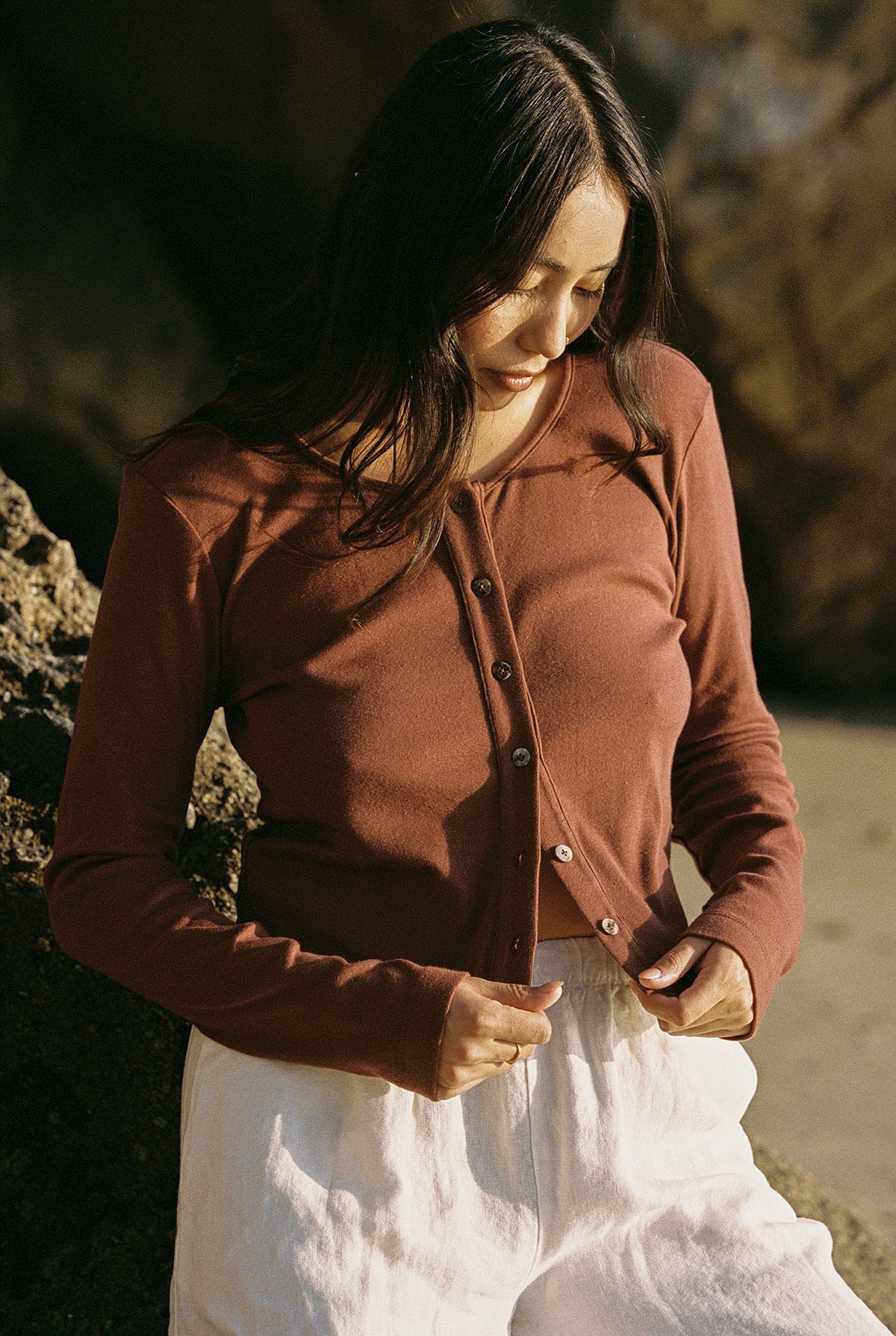 Mocha 
In a natural, rocky setting evoking a '90s closet vibe, a person with long hair stands outdoors. They wear the LA Relaxed Organic Rib Cardigan in brown and white pants, adjusting the buttons as they look down.