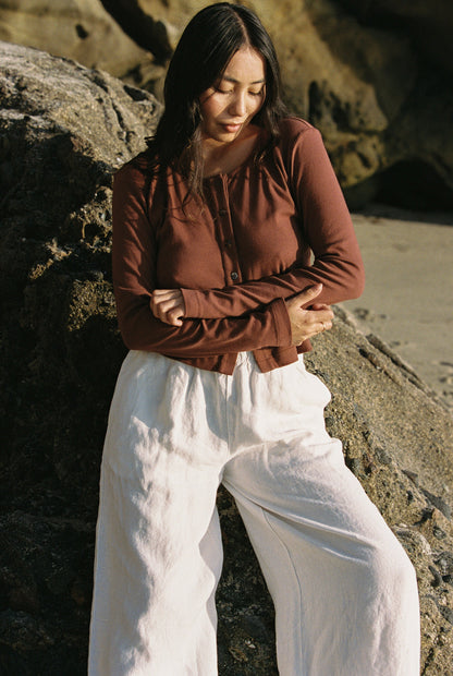 Mocha 
In a 90's Closet style, a person with long dark hair stands outdoors against a rock. They wear an LA Relaxed Organic Rib Cardigan in brown and white wide-leg pants made from organic cotton. The rocky setting is illuminated by natural light, possibly indicating a beach backdrop.