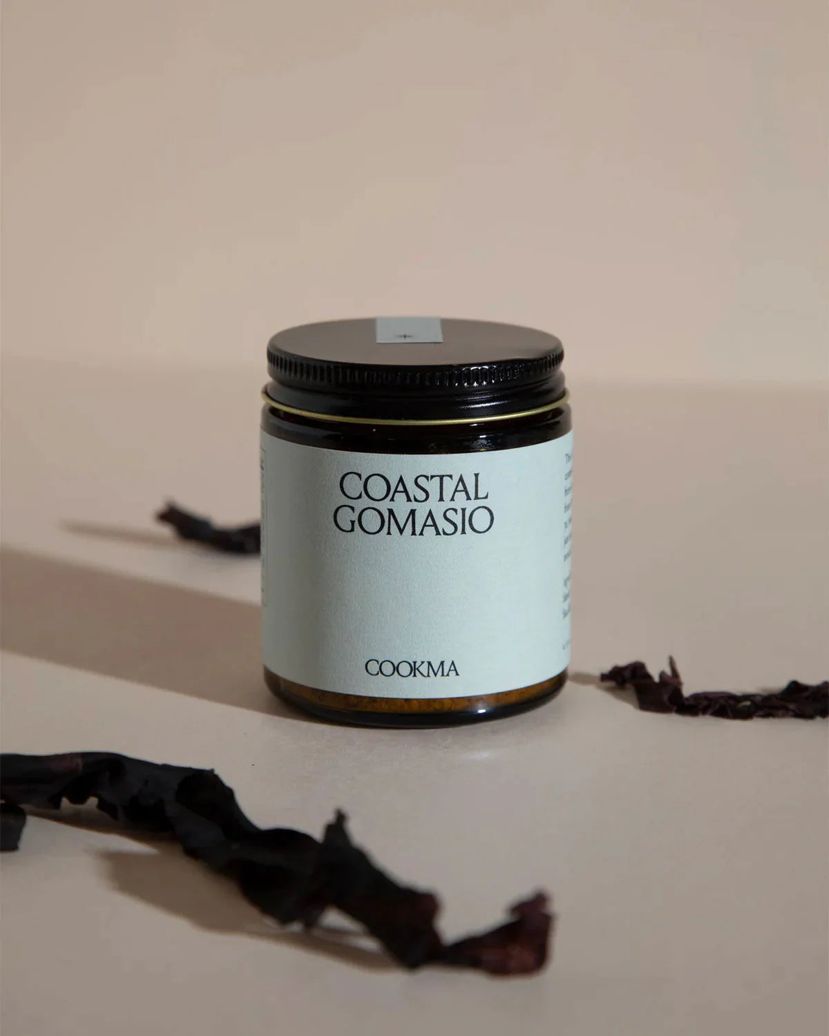 A jar from Cookma’s Mama Bundle sits on a beige surface with dark dried seaweed nearby—ideal for enriching postpartum meals and boosting nutrition with nutrient-rich flavor.