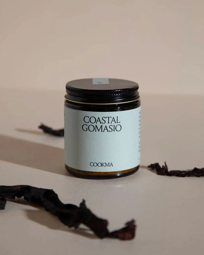 A jar from Cookma’s Mama Bundle sits on a beige surface with dark dried seaweed nearby—ideal for enriching postpartum meals and boosting nutrition with nutrient-rich flavor.