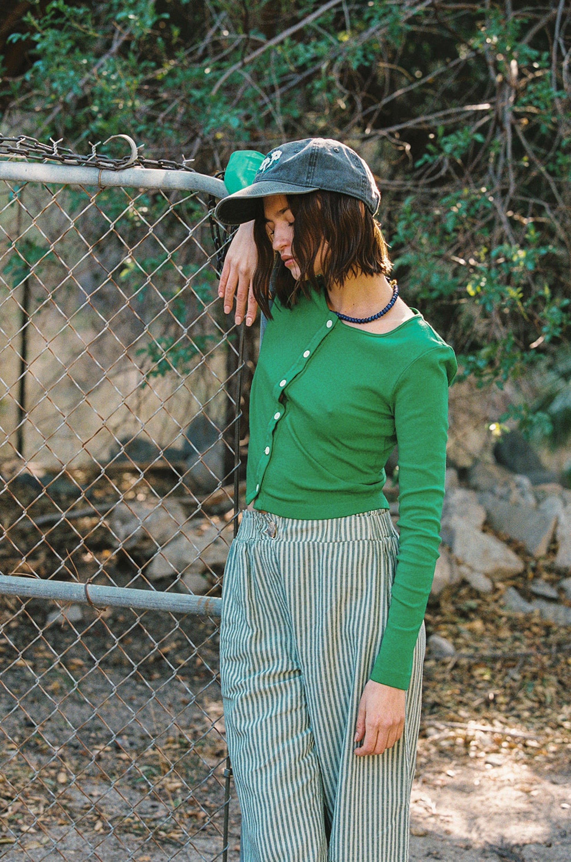 Grass
Someone wearing an Organic Rib Cardigan by LA Relaxed and striped pants leans on a chain-link fence outside. Sporting a dark hat with a '90s vibe, they exude relaxed style amidst greenery, effortlessly showcasing eco-friendly fashion made from organic cotton.