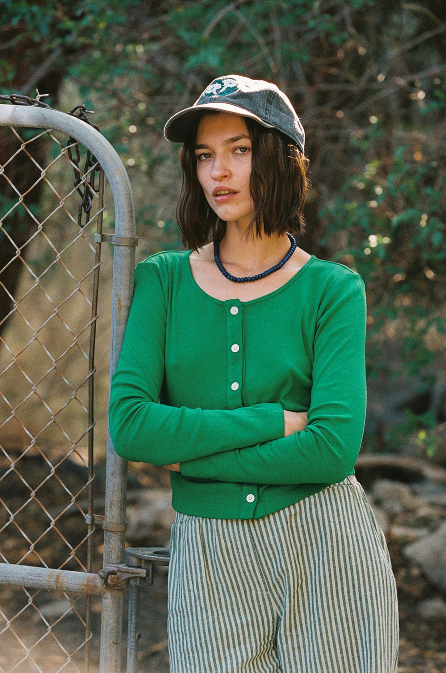 Grass 
A person stands with arms crossed by a wire fence outdoors, wearing an LA Relaxed Organic Rib Cardigan, striped pants, and a denim cap. The lush greenery and soft lighting create a 90's closet vibe in the background.