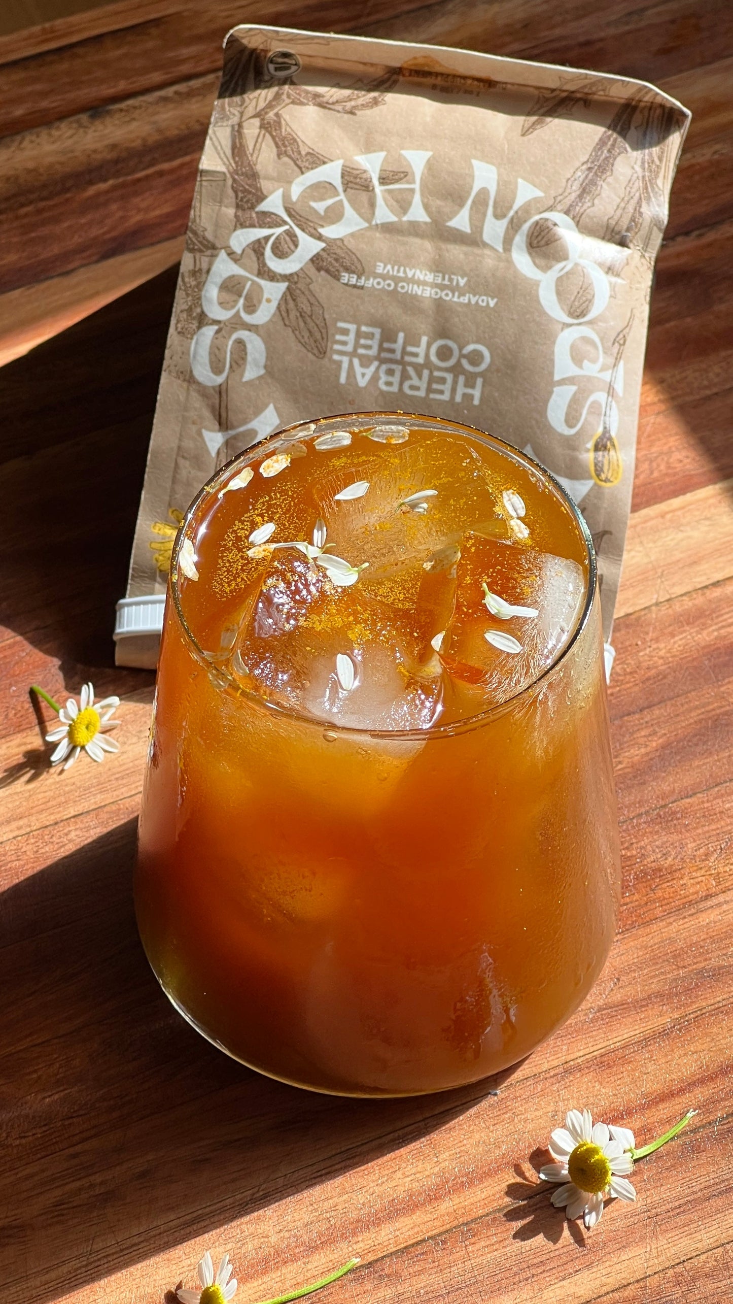 A glass of iced Herbal Coffee with ice cubes rests on a wooden table, garnished with small white flowers. Behind the drink, a partially visible brown packet labeled "WOODEN SPOON HERBS Herbal Coffee" adds to the scene. Sunlight casts shadows and highlights, creating a warm and inviting ambiance.