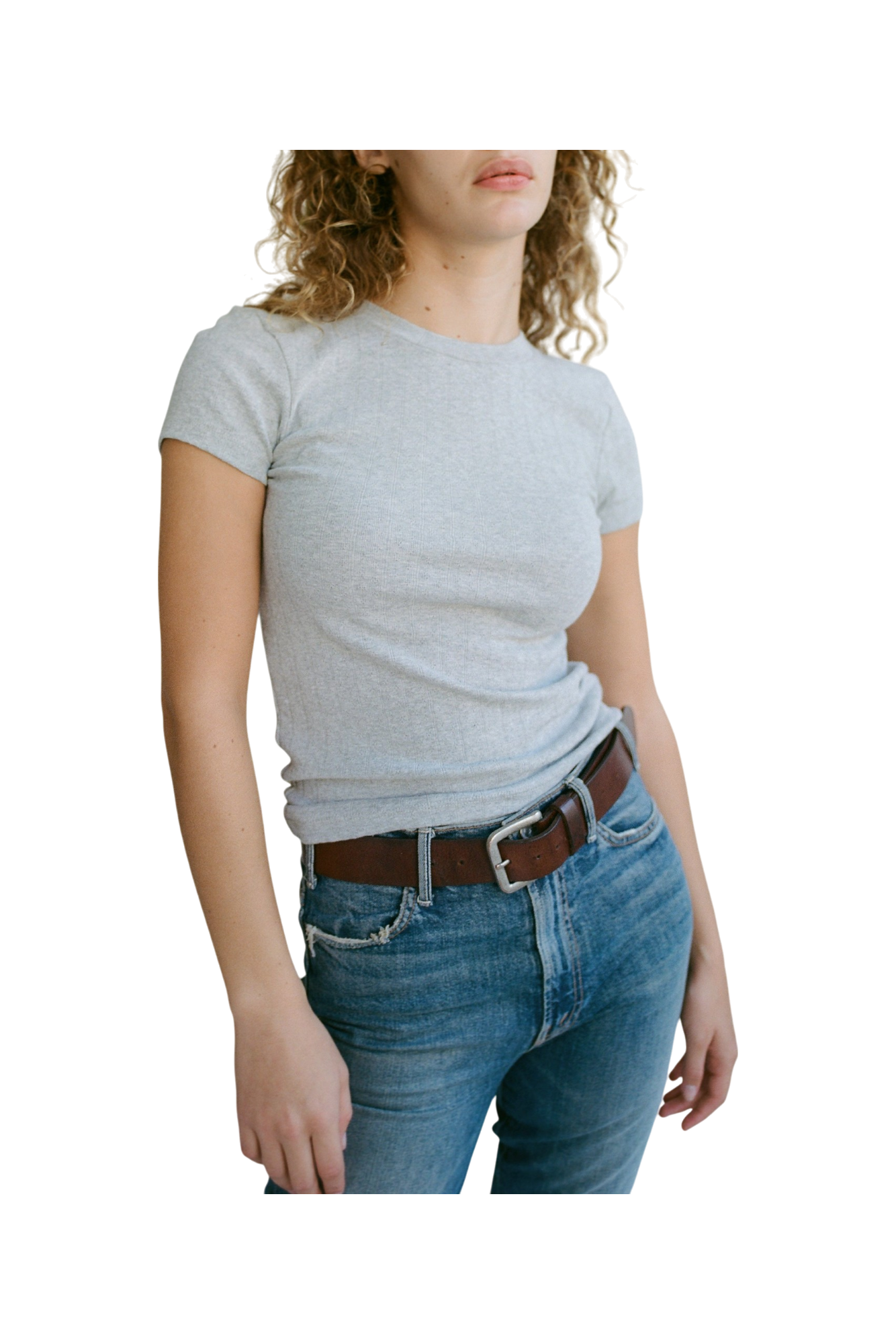 A person with curly hair is wearing an Organic Texture Tee in light gray by LA RELAXED, paired with blue jeans and a brown belt, standing with a relaxed posture against a black background.
