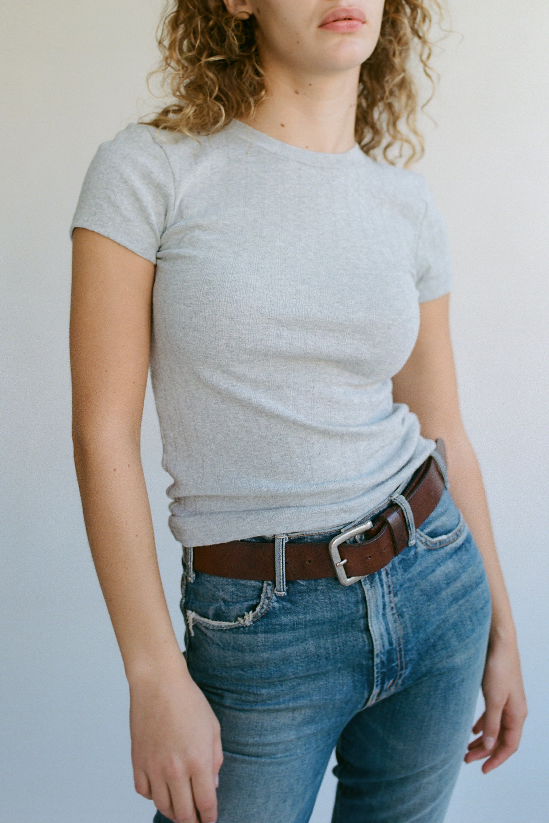 A person with curly hair wearing LA RELAXED's Organic Texture Tee in gray and blue jeans with a brown belt stands against a plain backdrop. The image highlights their upper body and casual outfit.