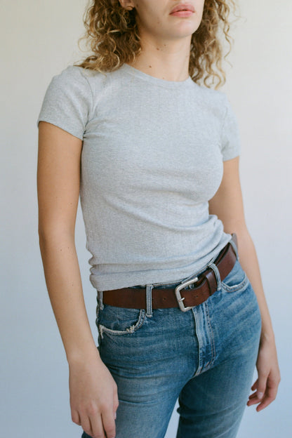 A person with curly hair wearing LA RELAXED's Organic Texture Tee in gray and blue jeans with a brown belt stands against a plain backdrop. The image highlights their upper body and casual outfit.