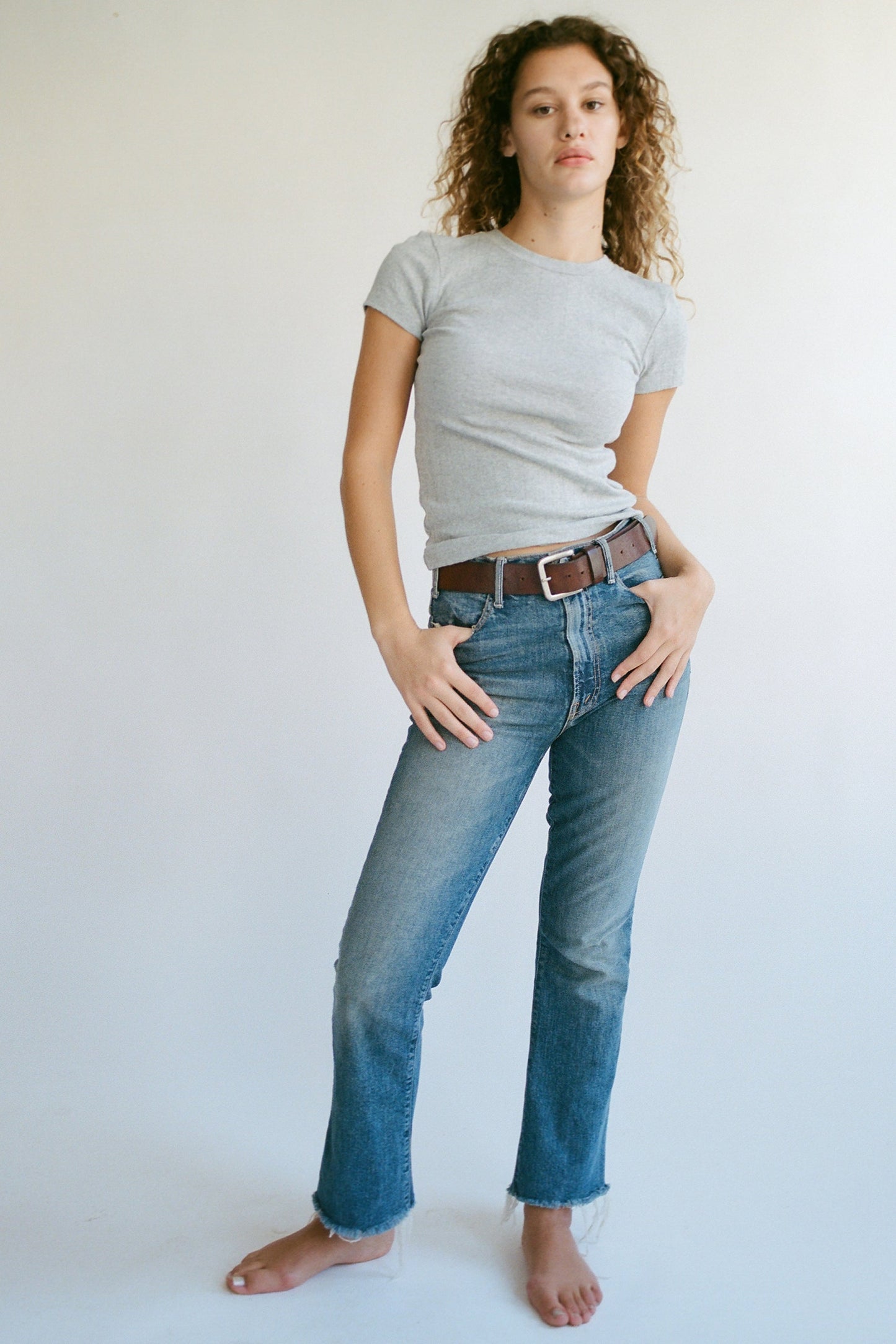 Against a plain backdrop, a person with curly hair stands barefoot in a fitted gray Organic Texture Tee by LA RELAXED, blue jeans, and brown belt. With one hand in their pocket and the other on their hip, they exude effortless style.