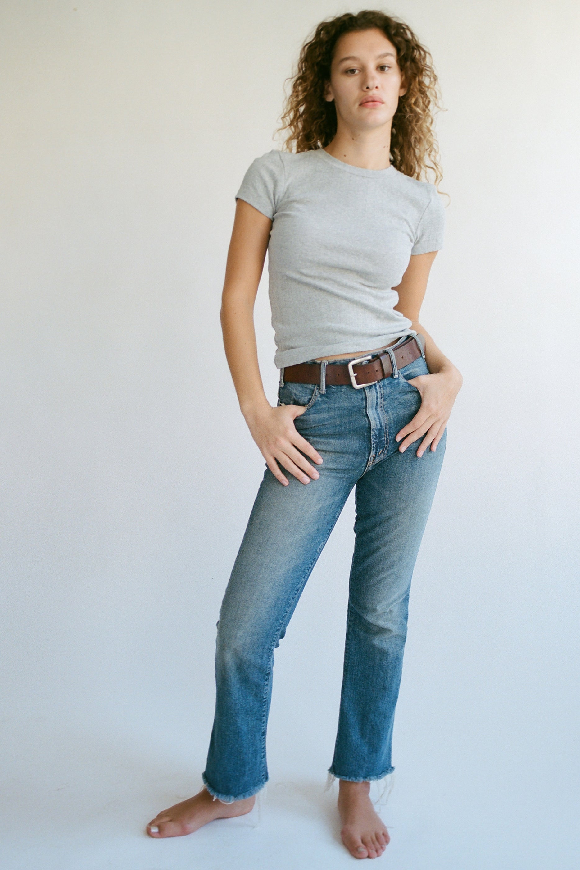 Against a plain backdrop, a person with curly hair stands barefoot in a fitted gray Organic Texture Tee by LA RELAXED, blue jeans, and brown belt. With one hand in their pocket and the other on their hip, they exude effortless style.
