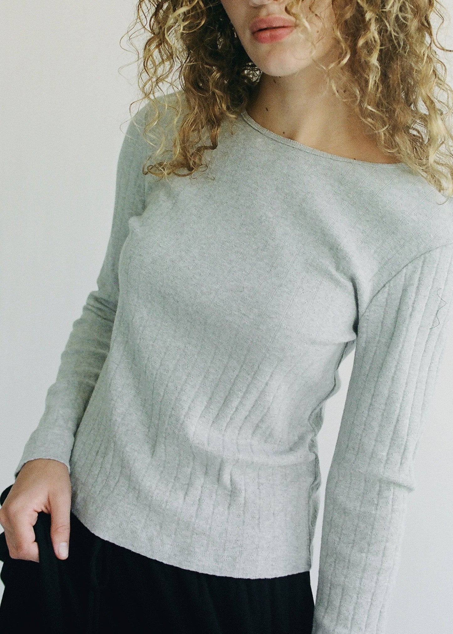A person with curly hair is wearing the Organic Texture Long Sleeve sweater by LA RELAXED in light gray, paired with black pants against a plain background, captured from the shoulders down.