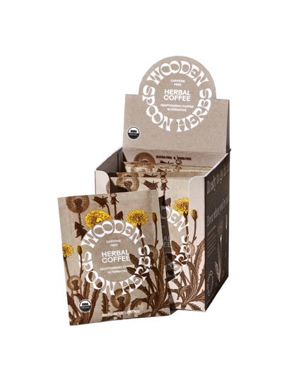 A product display box of WOODEN SPOON HERBS Herbal Coffee, highlighting a caffeine-free blend with multiple individual packets. The packaging features illustrations of dandelion and other plants in a natural earthy color scheme, with text indicating USDA organic certification and the inclusion of adaptogenic mushrooms.