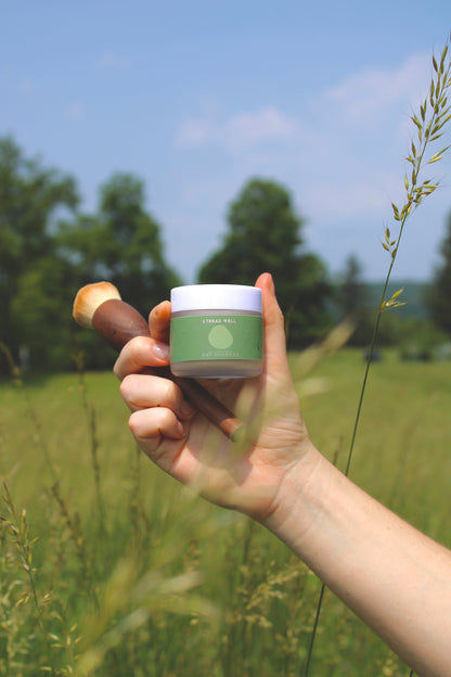 A hand holds a makeup brush and a green and white & Tread Well Dry Shampoo container in a grassy field with trees and blue sky, suggesting effortless hair volume in a natural, outdoor setting.