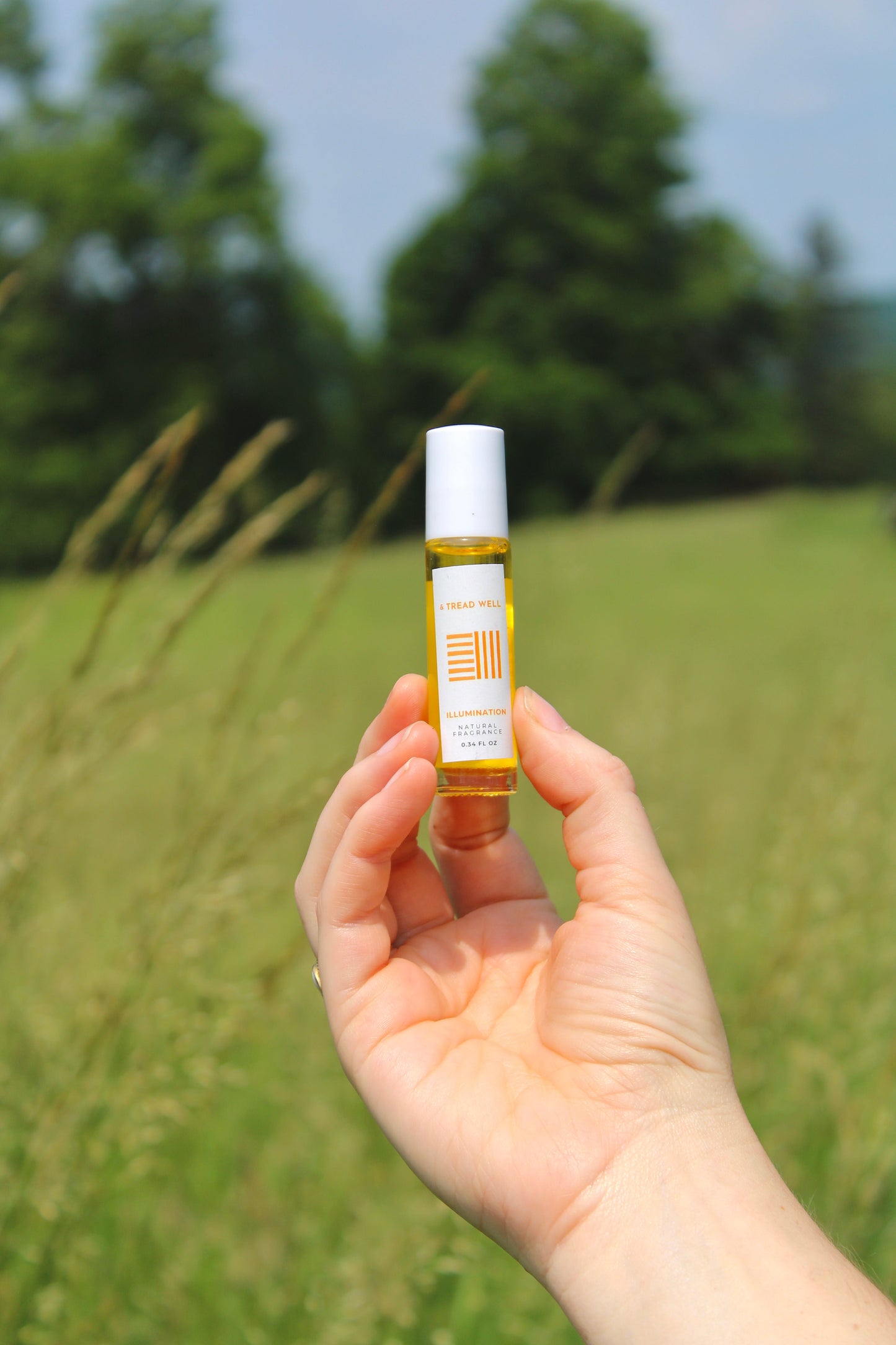 A hand holds & Tread Well’s Natural Fragrance – Illumination roll-on with a white cap and label, set against a grassy field and sunny sky, evoking natural aromatherapy and stress relief.