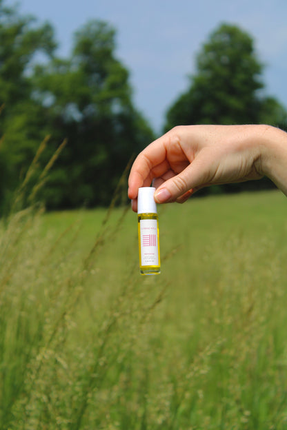 A hand holds a small bottle of & Tread Well Natural Fragrance – Meadow, its yellow liquid and white cap capturing the fresh, floral scent of geranium lavender outdoors beneath green trees and a blue sky.