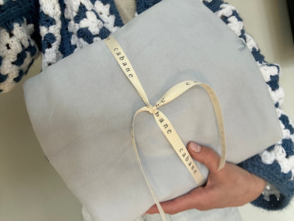 A person holds a folded, light gray fabric wrapped with a cream "cabane" ribbon. In the background, part of the BABY BLANKET by cabane childrenswear can be seen, featuring hand-quilted blue and white crocheted patterns that demonstrate exquisite craftsmanship.