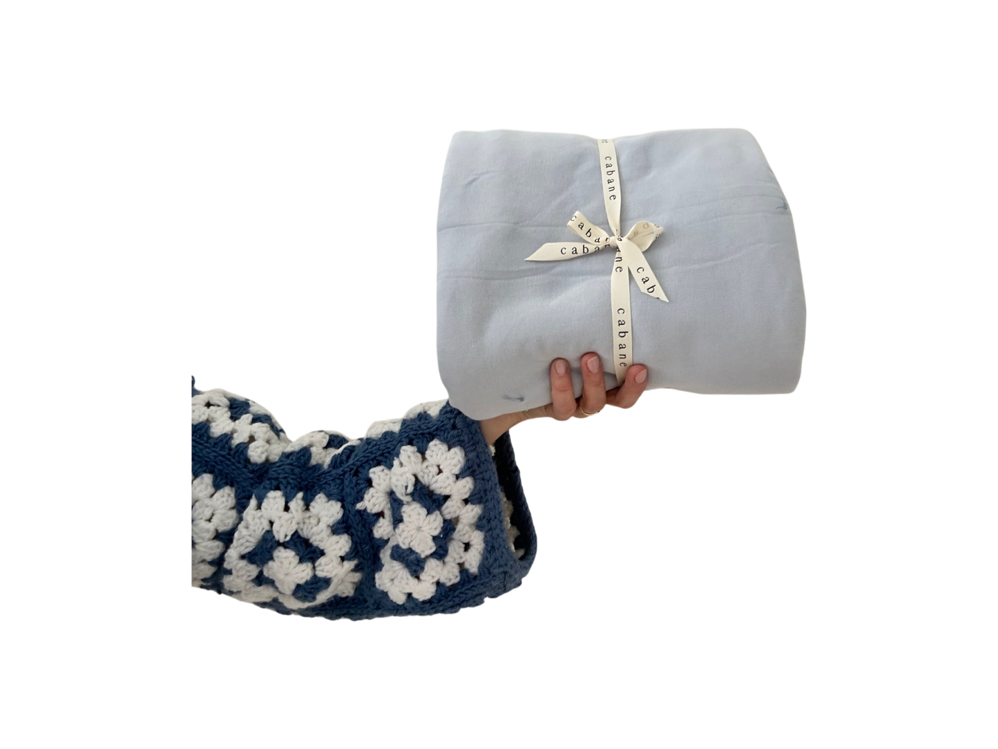 A hand in a blue and white crocheted sweater holds a neatly wrapped cabane childrenswear BABY BLANKET, indigo, tied with a ribbon against a black background.