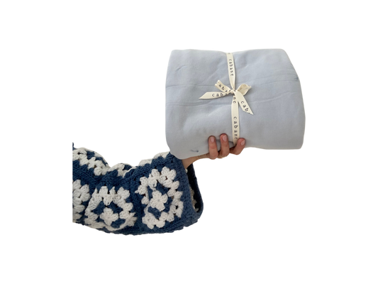 A hand in a blue and white crocheted sweater holds a neatly wrapped cabane childrenswear BABY BLANKET, indigo, tied with a ribbon against a black background.