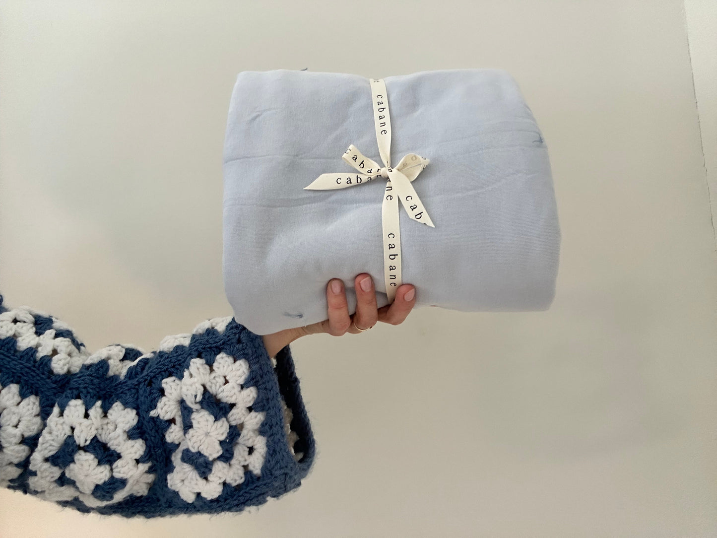A hand wears a blue and white crocheted sweater while holding a folded indigo BABY BLANKET from cabane childrenswear tied with a cream ribbon. The background is plain and light-colored.