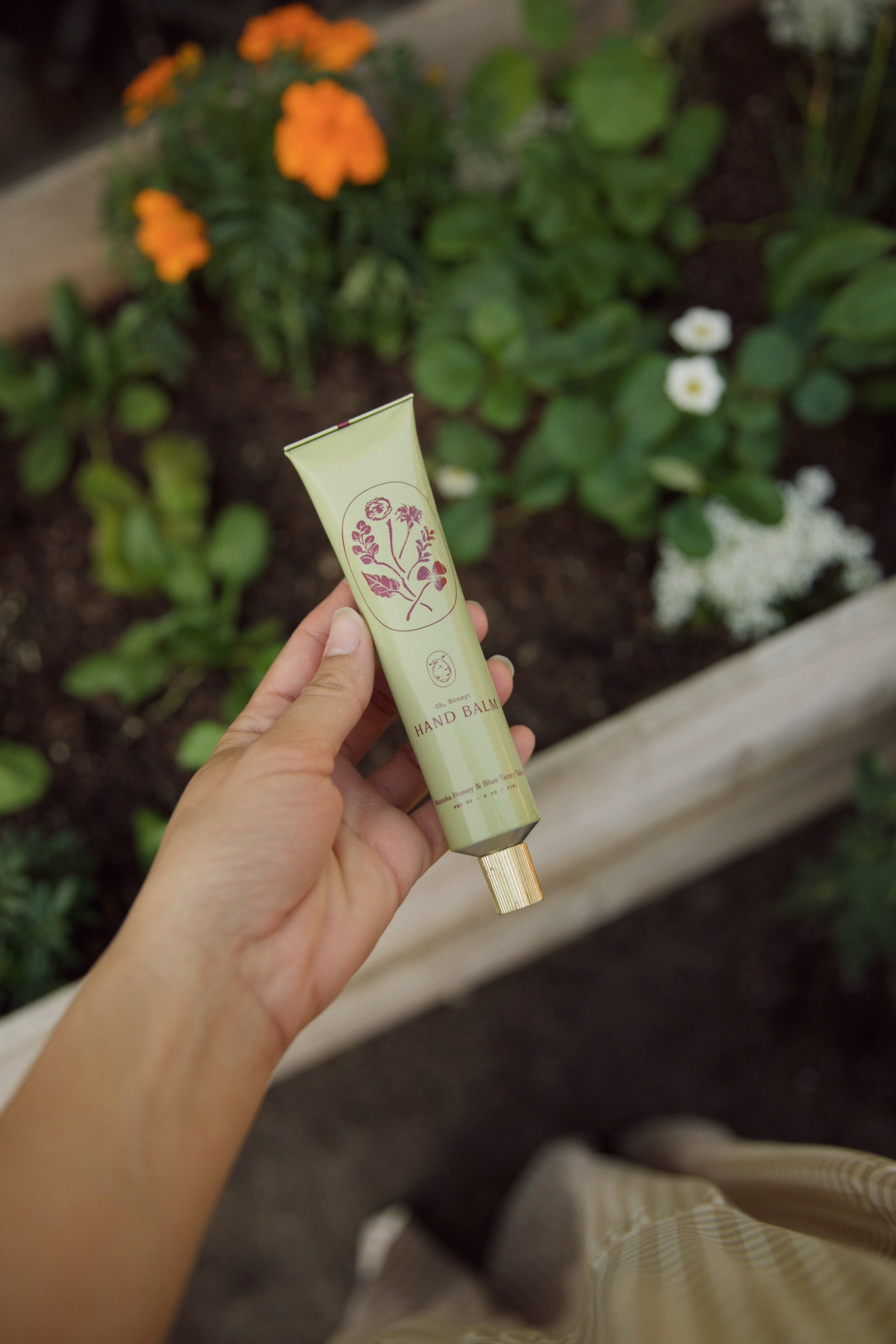 A hand holds a green tube of My Neighbor’s Tallow Manuka Honey & Blue Tansy Tallow Hand Cream with blue tansy floral designs over a garden bed filled with lush greenery and orange and white flowers.