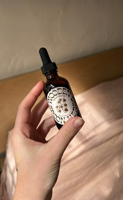 A hand holds an amber glass dropper bottle labeled "Head Helper Tincture" from Wooden Spoon Herbs. The label features an illustration of a tree, evoking a natural, herbal tincture vibe. The background is softly lit with a pink fabric and a wooden surface partially visible.