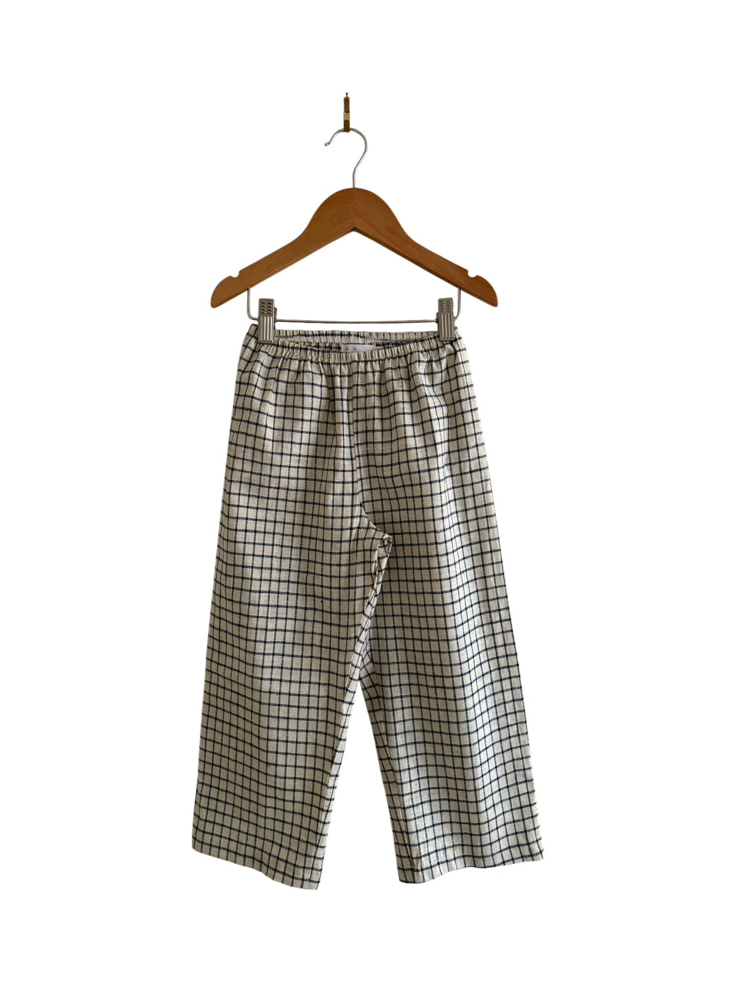 The PER SE PANT blue check by cabane childrenswear, featuring an elastic waistband and loose fit, is handmade in Vancouver. The pants hang on a wooden hanger against a black background.