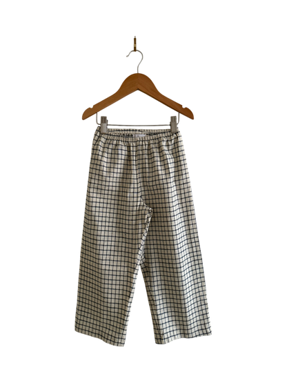 The PER SE PANT blue check by cabane childrenswear, featuring an elastic waistband and loose fit, is handmade in Vancouver. The pants hang on a wooden hanger against a black background.