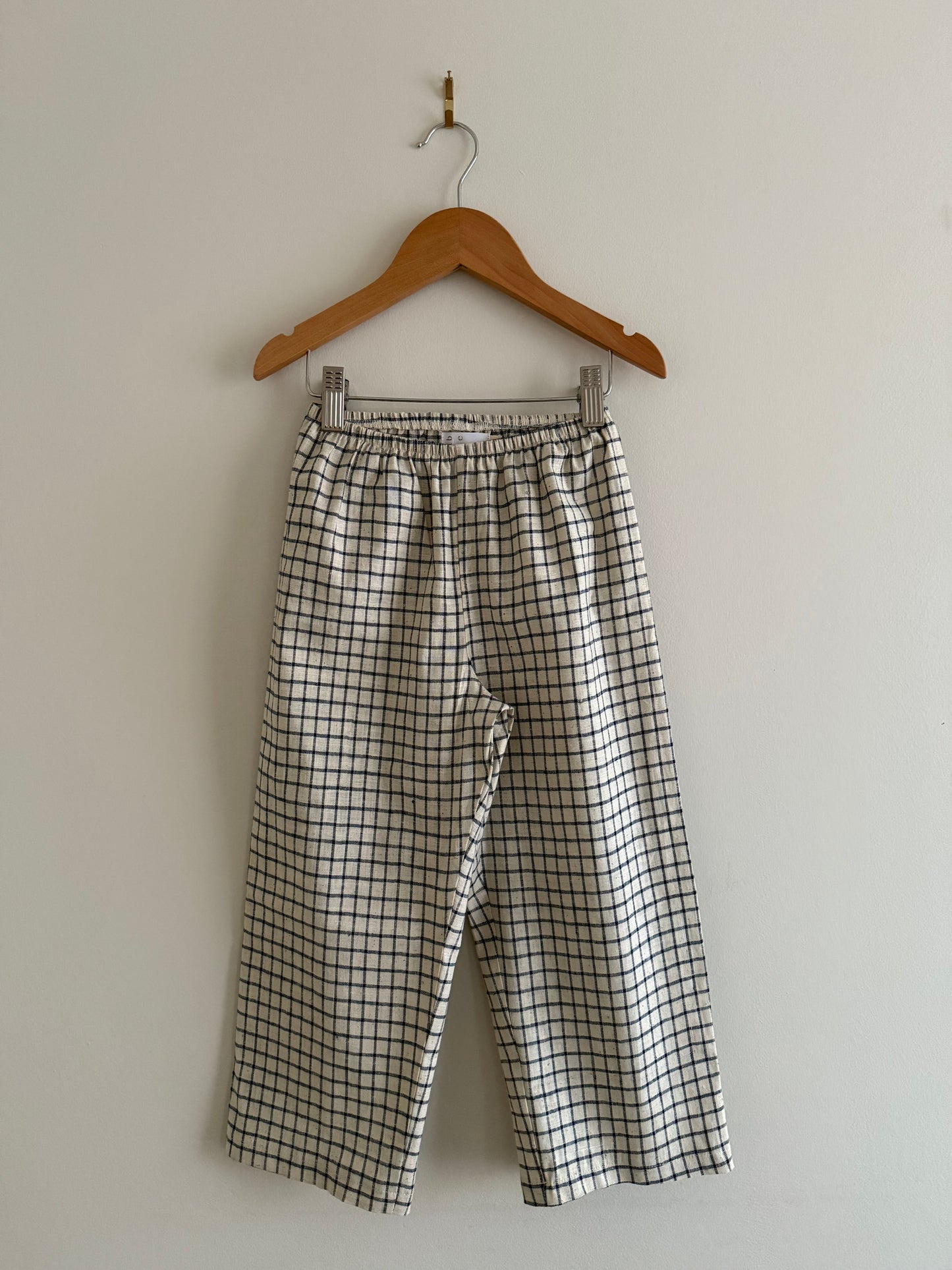 The PER SE PANT blue check by cabane childrenswear hangs on a wooden hanger against a plain white wall.