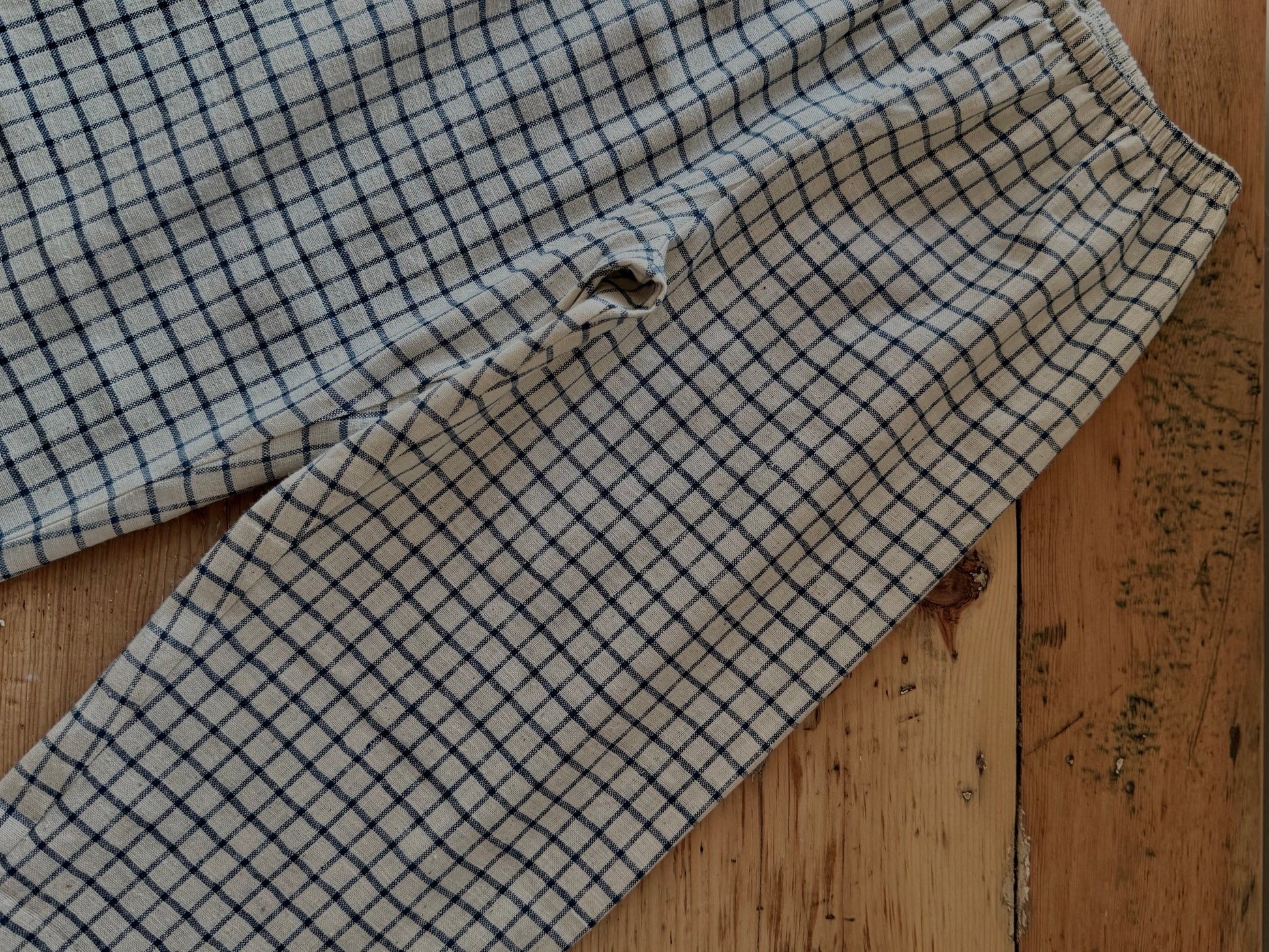 The PER SE PANT blue check by cabane childrenswear is a pair of light plaid pants made from organic cotton khadi with a blue check pattern, shown on wood, featuring a visible hole near one leg seam.