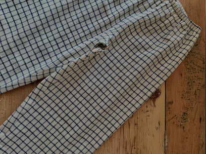 The PER SE PANT blue check by cabane childrenswear is a pair of light plaid pants made from organic cotton khadi with a blue check pattern, shown on wood, featuring a visible hole near one leg seam.