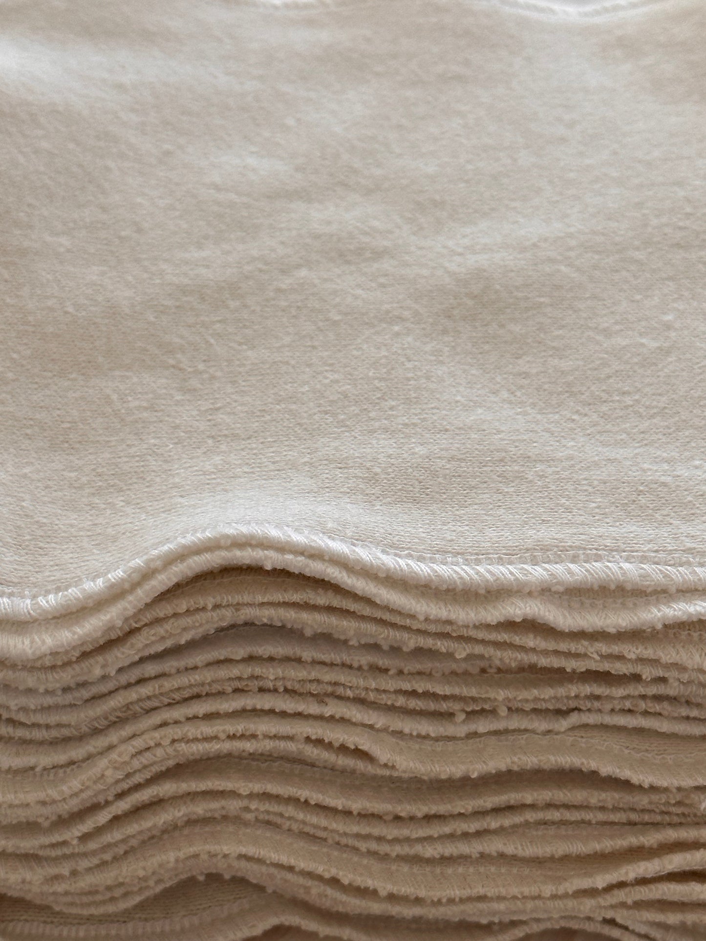 A close-up of a neat stack of cream-colored ORGANIC WASHCLOTHS (set of four) by cabane childrenswear, handmade in Vancouver from soft organic cotton with wavy edges and visible border stitching.
