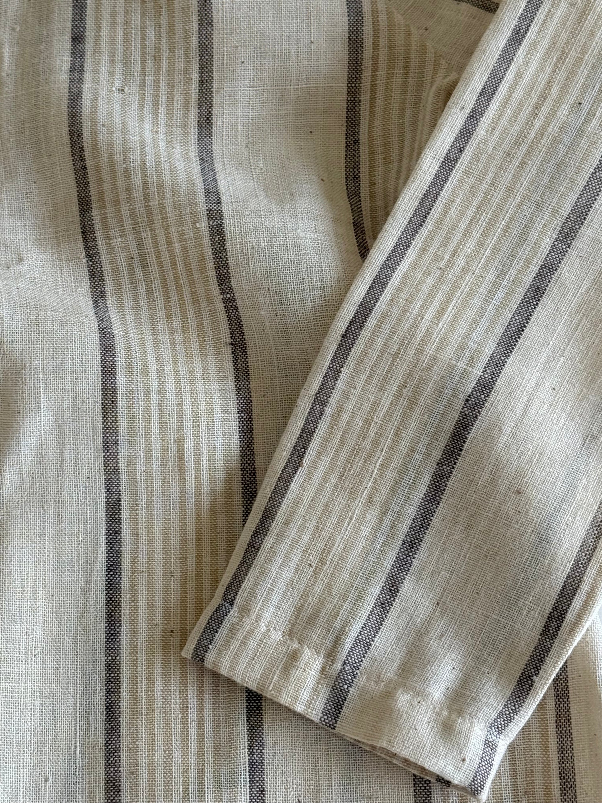 A close-up of the APRÈS WRAP natural stripe by cabane childrenswear shows breathable beige fabric with black and gray stripes, featuring a folded section that reveals the ethically handmade material.