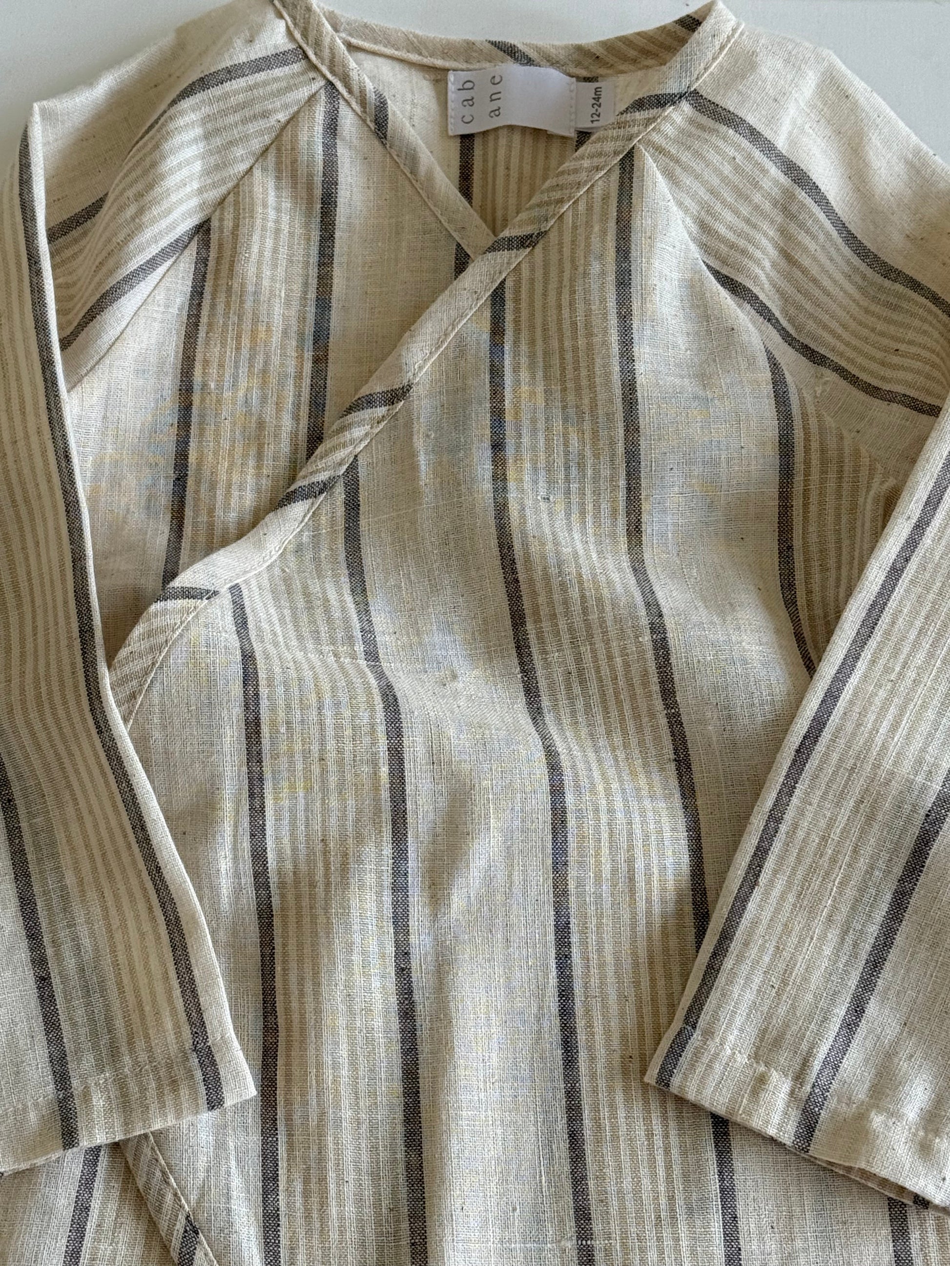 The APRÈS WRAP natural stripe by cabane childrenswear is a beige and cream long-sleeve linen top with vertical brown and gray stripes, shown flat on a light surface, featuring a visible label near the neckline.