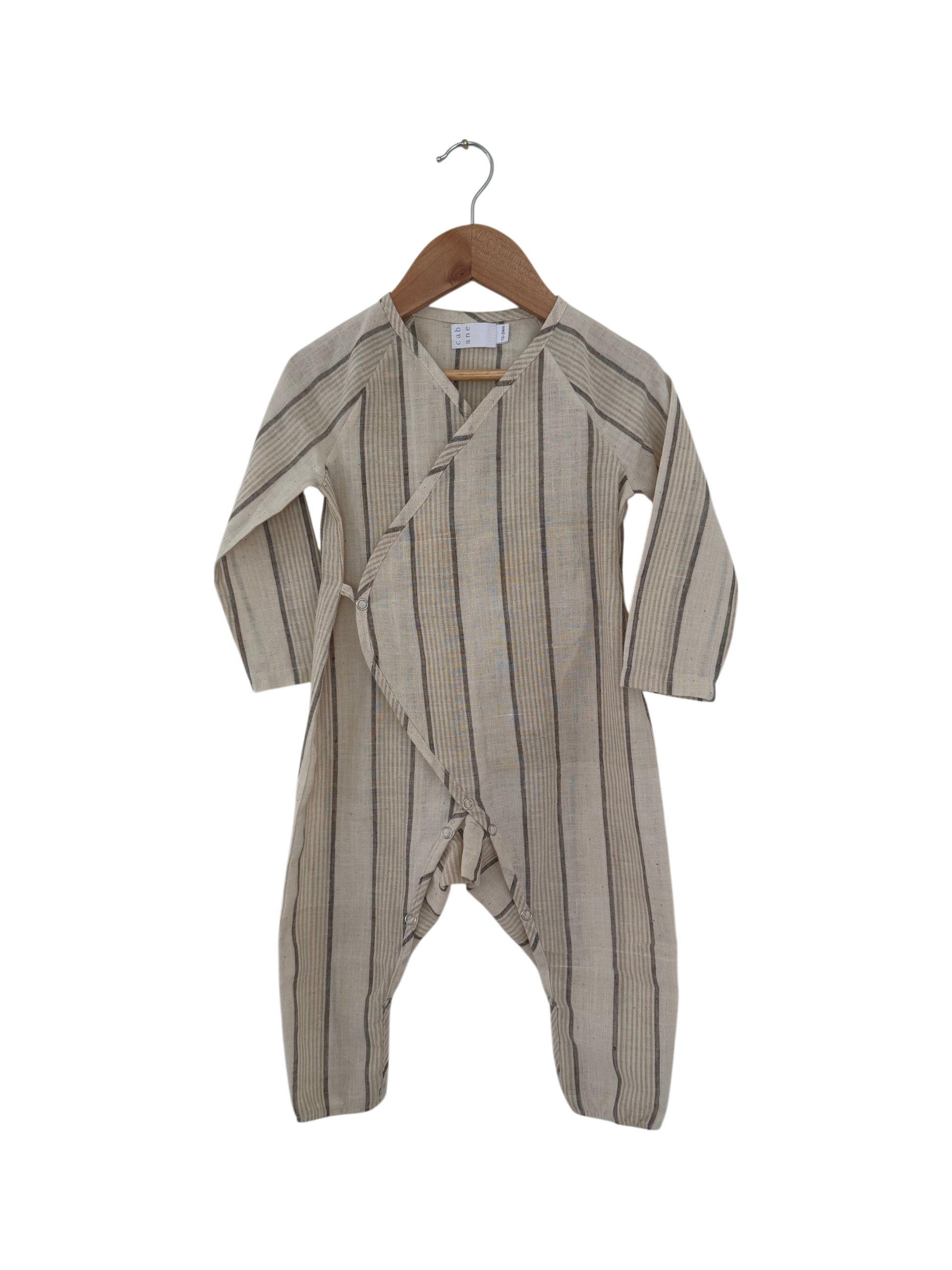 The cabane childrenswear APRÈS WRAP natural stripe is a beige, long-sleeved infant romper with thin black stripes, wrap-style front, and covered feet, shown on a wooden hanger against a black background.