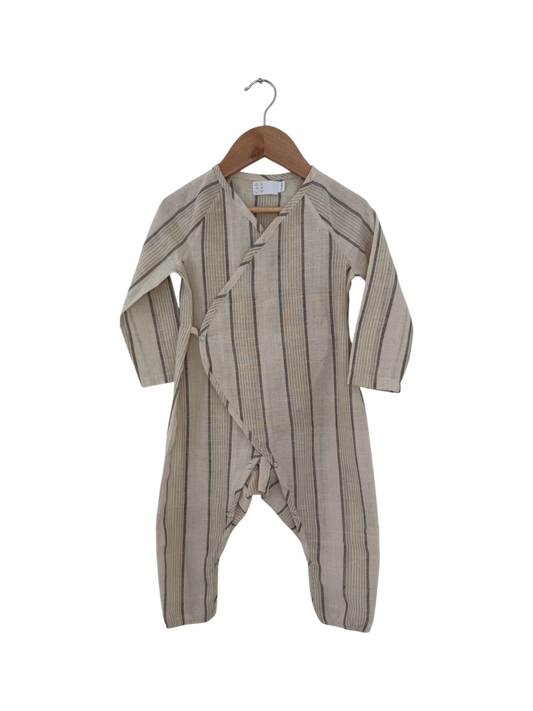 The cabane childrenswear APRÈS WRAP natural stripe is a beige, long-sleeved infant romper with thin black stripes, wrap-style front, and covered feet, shown on a wooden hanger against a black background.