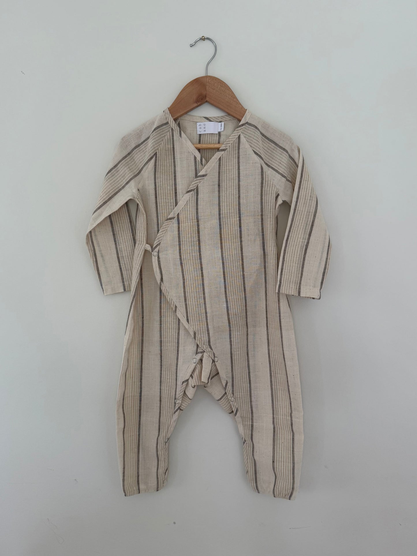 The APRÈS WRAP natural stripe by cabane childrenswear is a beige, long-sleeve baby romper with dark stripes and a side wrap with buttons, crafted from breathable fabric. Shown hanging on a wooden hanger against a white wall.