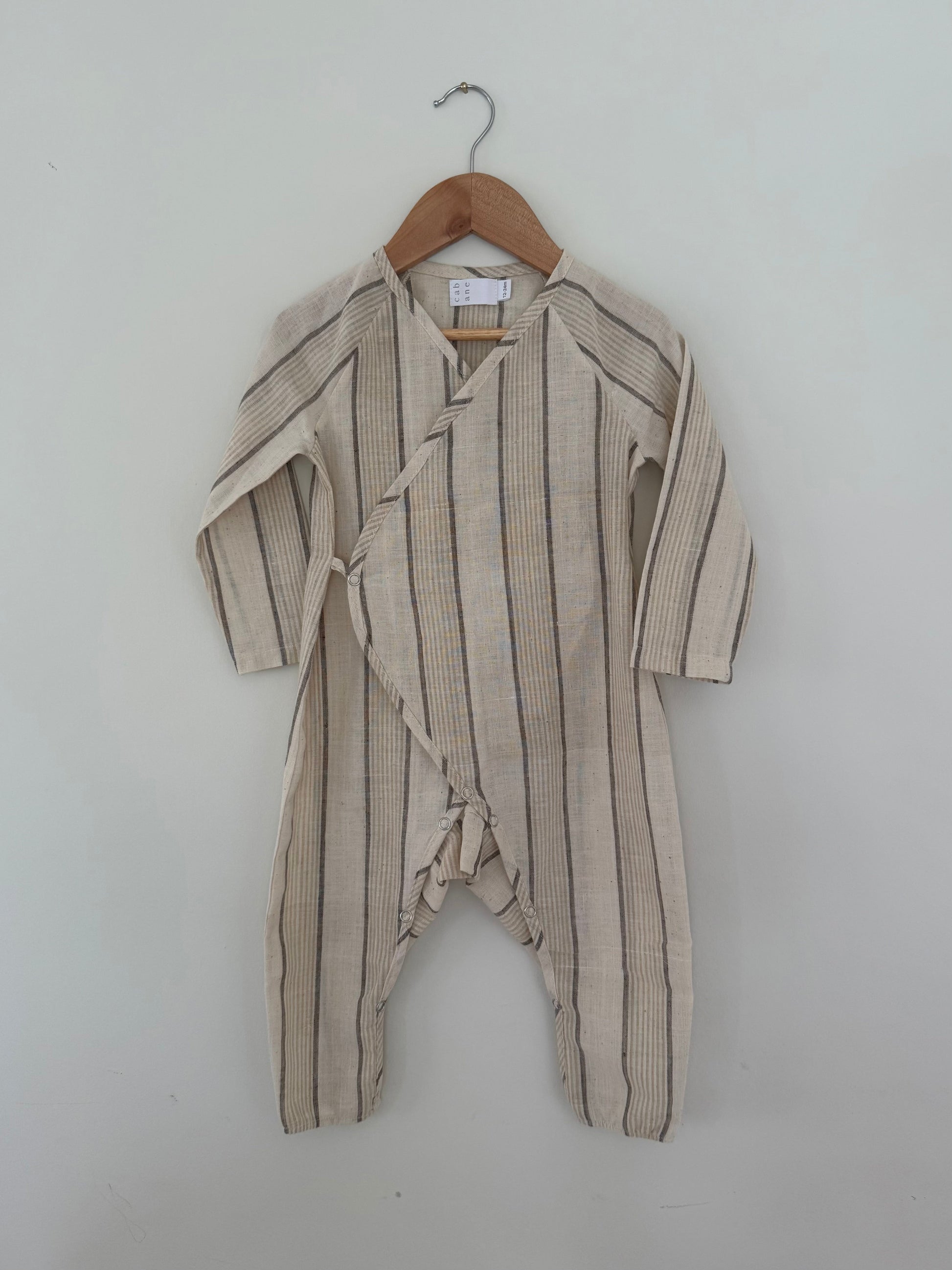 The APRÈS WRAP natural stripe by cabane childrenswear is a beige, long-sleeve baby romper with dark stripes and a side wrap with buttons, crafted from breathable fabric. Shown hanging on a wooden hanger against a white wall.