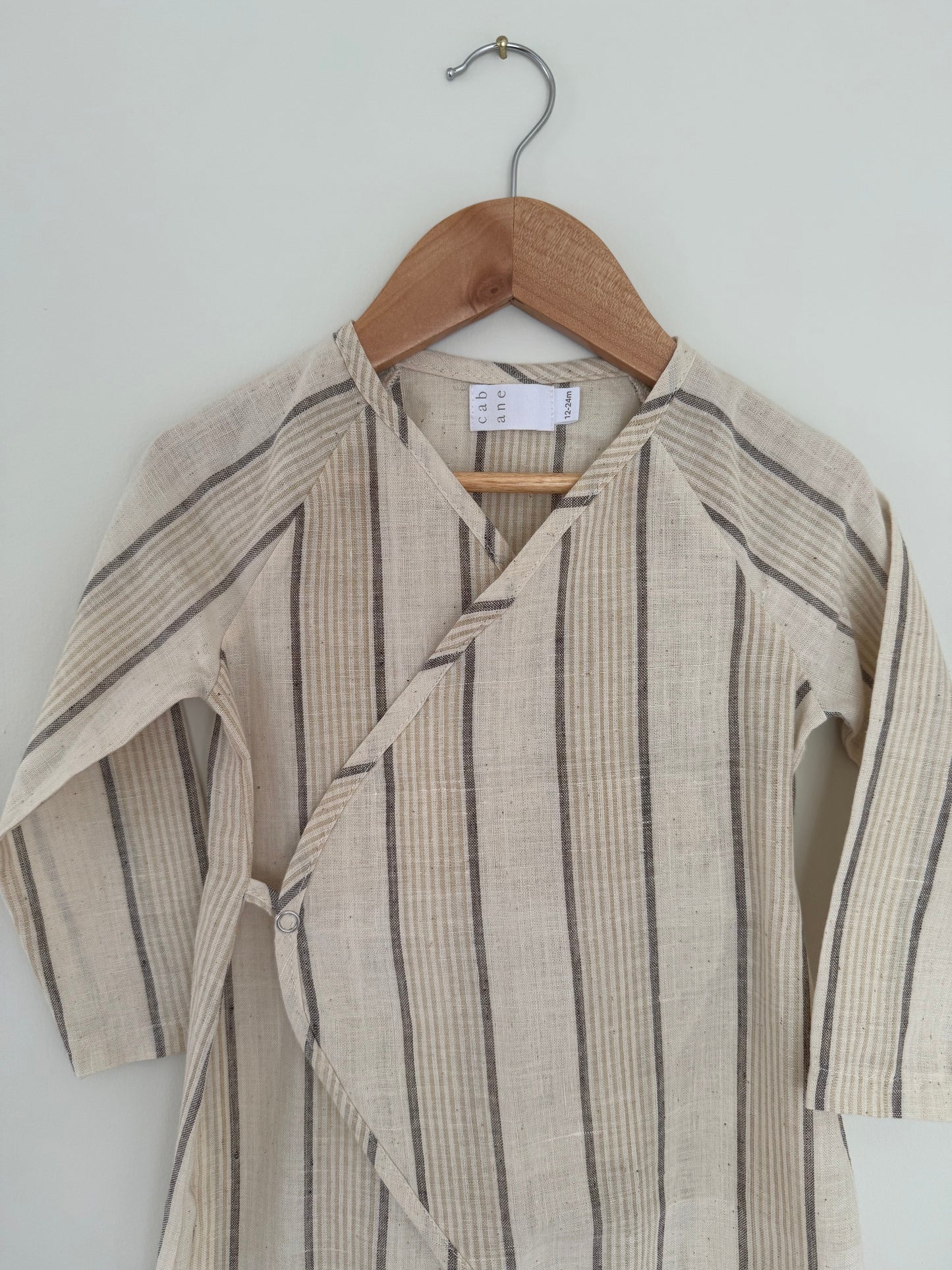 The APRÈS WRAP natural stripe by cabane childrenswear is a beige long-sleeve baby kimono with thin gray and tan stripes, made from breathable fabric, displayed on a wooden hanger against a light-colored wall.