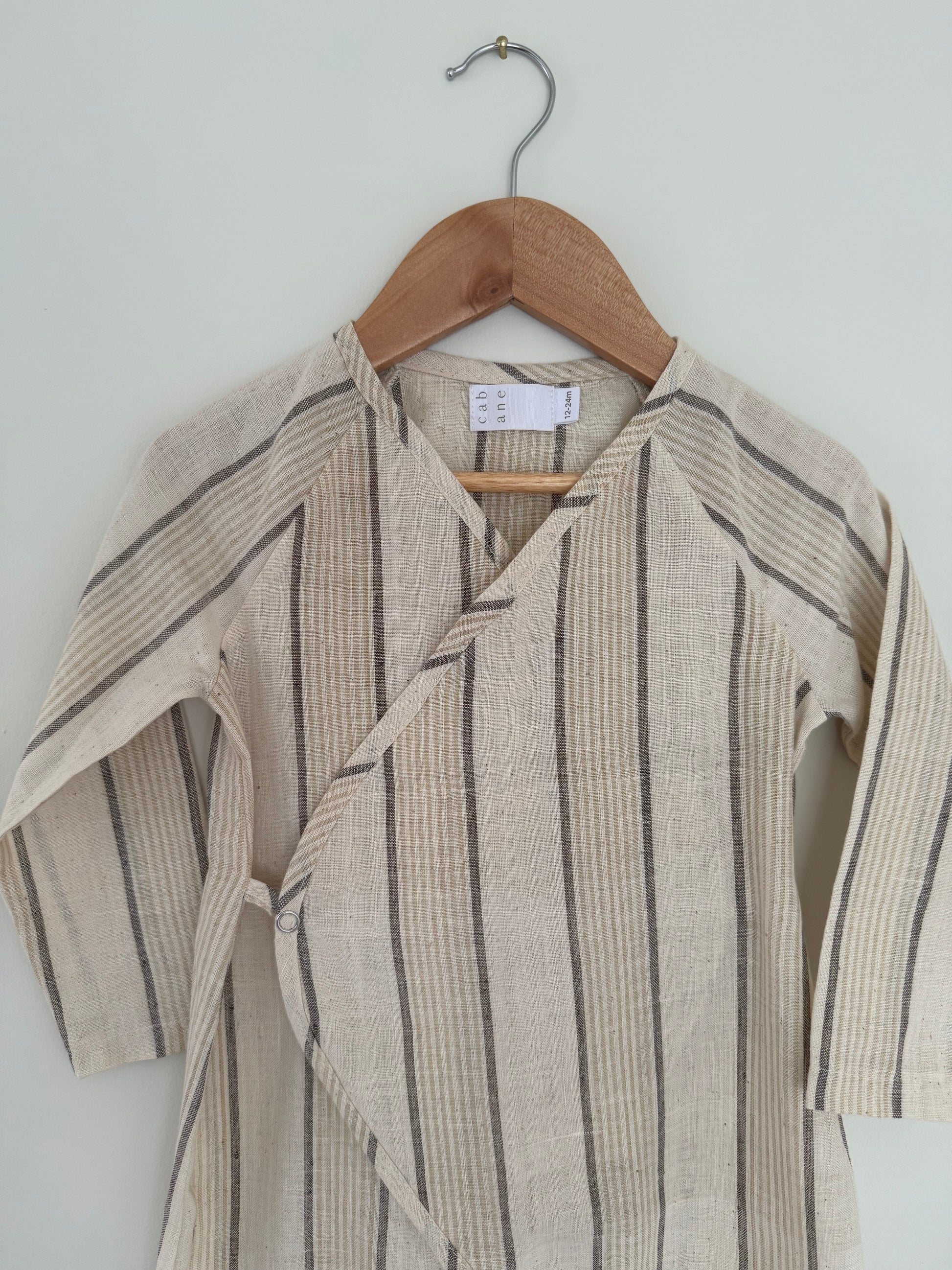 The APRÈS WRAP natural stripe by cabane childrenswear is a beige long-sleeve baby kimono with thin gray and tan stripes, made from breathable fabric, displayed on a wooden hanger against a light-colored wall.